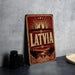 A vintage-style metal sign with the word "LATVIA" prominently displayed, featuring a cityscape and sunburst design.