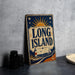 A vintage metal sign with the text "LONG ISLAND" and a lighthouse illustration, placed on a gray countertop next to some black camera lenses.