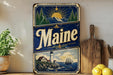 A vintage Maine-themed metal sign with a lighthouse, mountains, and a sun in the sky, displayed on a shelf next to a wooden cutting board and some lemons.