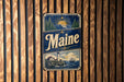 A vintage metal sign with the word "Maine" prominently displayed, featuring a lighthouse and a scenic coastal landscape.