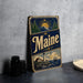 A vintage Maine-themed metal sign with a lighthouse, mountains, and a sun in the background, along with the word "Maine" prominently displayed.