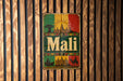 A vintage metal sign with the word "Mali" displayed prominently in the center, surrounded by various cultural and architectural elements such as a sun, buildings, and a camel.