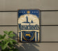 A vintage metal sign with the text "Massachusetts" displayed prominently, featuring a lighthouse and sailboat illustration. The sign is mounted on a wall, and there is a plant visible to the left of the sign.