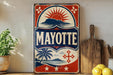 A vintage-style metal sign with the word "MAYOTTE" prominently displayed, featuring a sun and palm tree design. The sign is placed on a shelf next to a wooden cutting board and a potted plant.