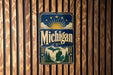 A vintage metal sign with the word "Michigan" prominently displayed, featuring a sun and clouds design, is mounted on a wooden wall.