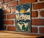A vintage-style metal sign with the word "Michigan" prominently displayed, featuring a sun and clouds design, is mounted on a brick wall.