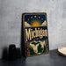 A vintage-style metal sign with the word "Michigan" prominently displayed, featuring a sun and clouds design, and a maple leaf illustration. The sign is placed on a gray countertop next to some black camera lenses.