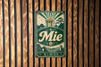 A vintage metal sign with the word "Mie" prominently displayed, featuring a sun and pagoda-like structure, is mounted on a wooden wall.