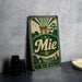 A vintage metal sign with a green and gold design, featuring a Japanese-style torii gate and mountains, along with the word "Mie" prominently displayed.