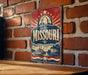 A vintage-style metal sign with the text "Missouri" prominently displayed, featuring a riverboat illustration and a city skyline in the background.