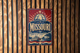 A vintage-style metal sign with the word "Missouri" prominently displayed, featuring a riverboat and a city skyline.