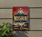 A vintage-style metal sign with the word "Missouri" prominently displayed, featuring a riverboat and a city skyline in the background.