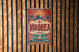 A vintage metal sign with a tropical design, featuring palm trees, mountains, and a boat, is mounted on a wooden wall.