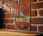 A vintage metal sign with the word "MOROCCO" prominently displayed, featuring a star and decorative elements, is mounted on a brick wall.