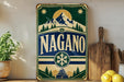 A vintage-style sign with the word "NAGANO" prominently displayed, featuring a mountain landscape and snowflake design. The sign is placed on a wooden surface, with a potted plant, a wooden cutting board, and some lemons nearby.