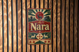A vintage-style metal sign with the name "Nara" displayed prominently in the center, surrounded by various decorative elements such as a sunburst design, a deer, and other traditional Japanese motifs.