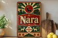 A vintage-style sign with a red and green color scheme, featuring a deer and pagoda in the background, along with the word "Nara" prominently displayed. The sign is placed on a wooden surface, with a potted plant and a wooden cutting board nearby.