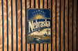 A metal sign with the word "Nebraska" on it, featuring a sun and windmill design, is displayed on a wooden wall.