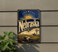 A vintage-style metal sign with the word "Nebraska" prominently displayed, featuring a sun and windmill illustration. The sign is mounted on a wall, and there is a plant visible to the left of the sign.
