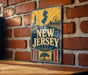 A vintage-style metal sign with the text "NEW JERSEY" prominently displayed, featuring a lighthouse, ferris wheel, and tree silhouette.