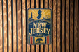 A vintage-style metal sign with the text "NEW JERSEY" prominently displayed, featuring a lighthouse, ferris wheel, and other nautical elements.
