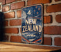 A vintage-style sign with the text "NEW ZEALAND" prominently displayed, featuring a kiwi bird and a mountain landscape. The sign is mounted on a brick wall, and there is a wooden table in the foreground.