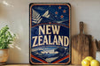 A vintage-style sign for New Zealand, featuring a mountain landscape, stars, and a kiwi bird, is displayed on a shelf next to a wooden cutting board and some lemons.