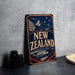 A vintage-style metal sign with the text "NEW ZEALAND" prominently displayed, featuring a mountain landscape and stars, is leaning against a concrete wall.