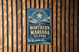 A vintage-style sign for the Northern Mariana Islands, featuring a blue background with a white star and palm trees, mounted on a wooden wall.