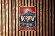 A vintage-style metal sign with the word "NORWAY" prominently displayed, featuring a mountain landscape and a ship, is mounted on a wooden wall.