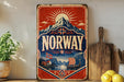 a vintage-style sign with the word "NORWAY" prominently displayed, surrounded by a scenic illustration of mountains, a ship, and a sun. The sign is placed on a shelf next to a wooden cutting board and some potted plants, creating a rustic and charming atmosphere.