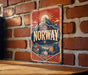 A vintage metal sign with the word "NORWAY" prominently displayed, featuring a mountain landscape and a Viking ship, mounted on a brick wall.
