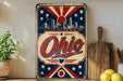 A vintage-style metal sign with the word "Ohio" prominently displayed, featuring a city skyline and stars in the background. The sign is placed on a shelf next to a wooden cutting board and a potted plant.