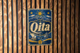 A vintage-style metal sign with the word "Qita" displayed prominently in the center, surrounded by various decorative elements such as mountains, sun, and flowers.