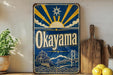 a vintage-style metal sign with the word "Okayama" prominently displayed, along with a sun and bridge illustration. The sign is placed on a shelf next to a potted plant and a wooden cutting board, creating a rustic and charming atmosphere.