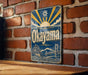 A vintage metal sign with the word "Okayama" displayed prominently, featuring a sun and bridge in the background.