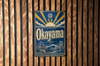 A vintage metal sign with the word "Okayama" displayed prominently, featuring a sun and clouds design, mounted on a wooden wall.