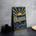 A vintage-style metal sign with the word "Okayama" prominently displayed, featuring a sun and clouds design, along with a castle and bridge in the background.