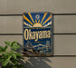 A vintage-style sign for "Okayama" is displayed on a wall, featuring a sun and clouds design with a bridge and castle in the background.