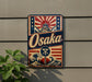 A vintage-style metal sign with the word "Osaka" prominently displayed, featuring a wave and a building with a Japanese-style roof.