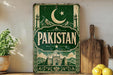 a vintage-style sign with the word "PAKISTAN" prominently displayed, surrounded by a green and white color scheme with a crescent moon and star motif. The sign is placed on a wooden surface, with a potted plant, a wooden cutting board, and some lemons nearby.