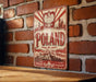 A vintage-style metal sign with the word "POLAND" prominently displayed, featuring a red and white color scheme and an illustration of a castle or palace. The sign is mounted on a brick wall, and there is a wooden table in the foreground.