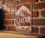 A vintage-style sign with the word "Qatar" prominently displayed, featuring a camel and a desert landscape, mounted on a brick wall.