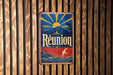 A vintage metal sign with a tropical landscape and sun design, mounted on a wooden wall.
