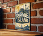 A vintage-style metal sign with the text "RHODE ISLAND" and a lighthouse illustration is displayed on a brick wall.