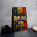 a vintage-style metal sign with the word "ROMANIA" prominently displayed, along with a castle and sun motif. The sign is placed on a gray surface, with a stack of black camera lenses and a white plate visible in the background.