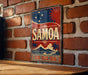 A vintage-style sign with the word "SAMOA" prominently displayed, featuring a red, white, and blue color scheme with a sun and stars design. The sign is mounted on a brick wall, and there is a wooden table below it.