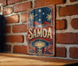 A vintage-style metal sign with the word "SAMOA" prominently displayed, featuring a tropical landscape with palm trees, a sunset, and stars.