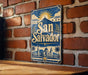 A vintage-style sign for San Salvador, featuring a sun and palm trees, is displayed on a brick wall.