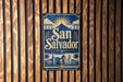 A vintage-style sign for San Salvador, featuring a sun and palm trees, is displayed on a wooden wall.